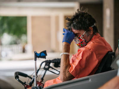 A man in a mask and gloves rests his head on his hand while seated in a wheelchair