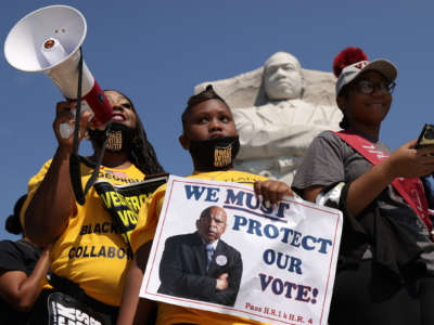 A girl in a yellow shirt holds a sign reading "PROTECT OUR RIGHT TO VOTE" during an outdoor protest