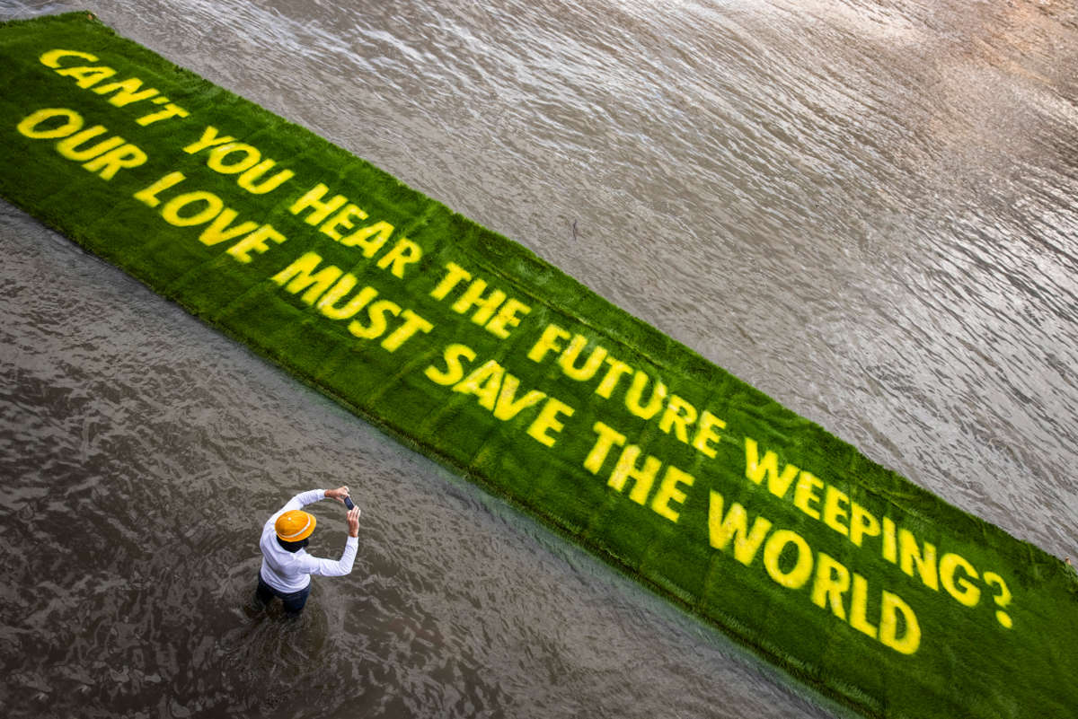 A person photographs a large banner of sod reading "CAN YOU HEAR THE PEOPLE WEEPING? OUT LOVE MUST SAVE THE WORLD" as it floats into a river