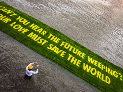 A person photographs a large banner of sod reading "CAN YOU HEAR THE PEOPLE WEEPING? OUT LOVE MUST SAVE THE WORLD" as it floats into a river