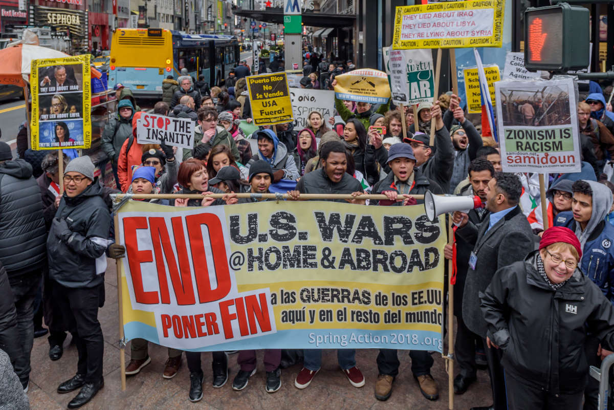 People chant behind a banner reading "END US WARS AT HOME AND ABROAD" in both english and spanish during a pre-covid rally