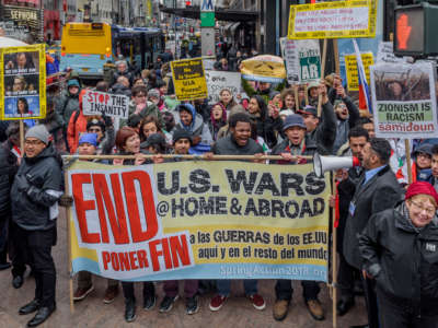 People chant behind a banner reading "END US WARS AT HOME AND ABROAD" in both english and spanish during a pre-covid rally