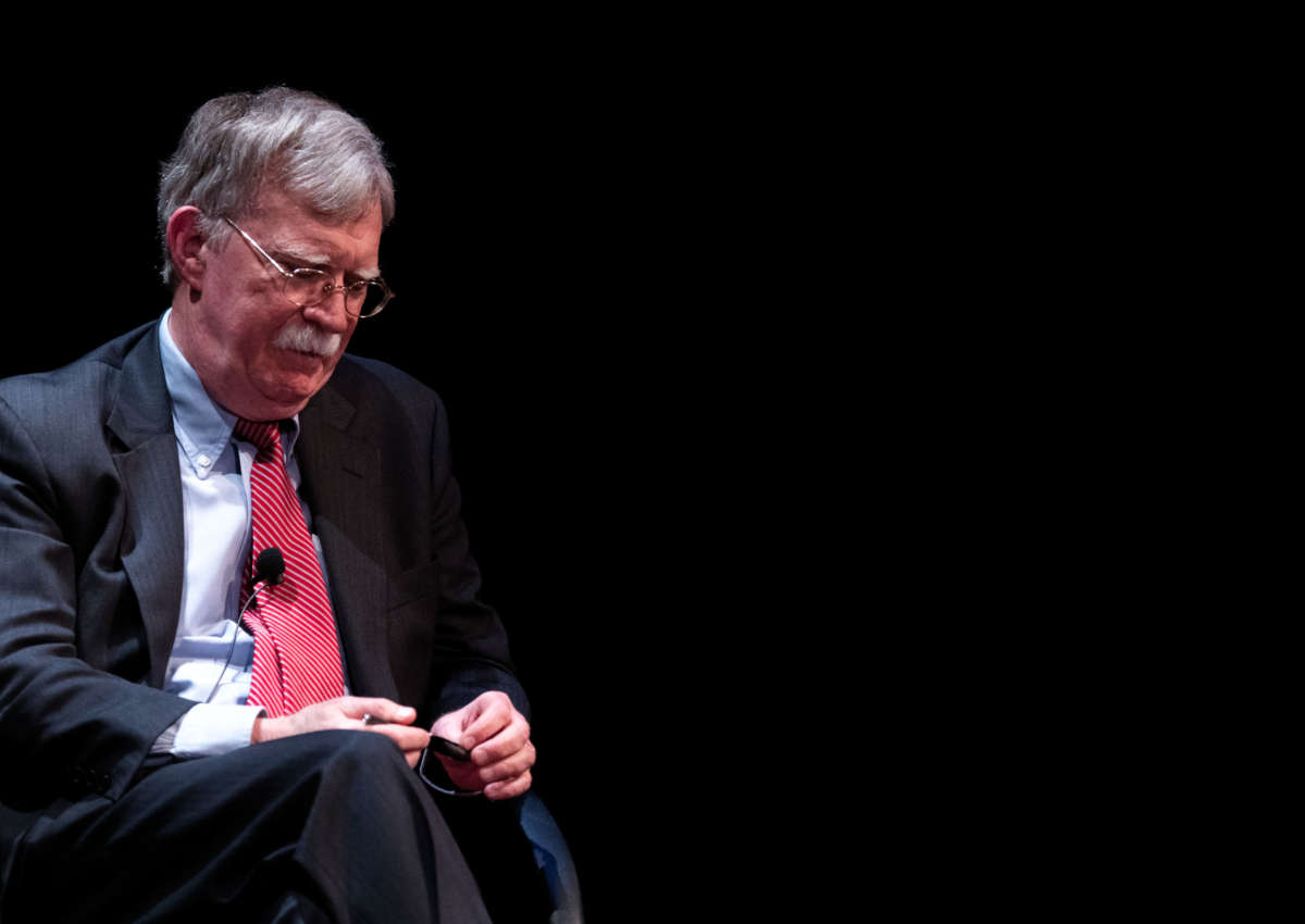 John Bolton sits on an otherwise dark stage looking like a skinny walrus