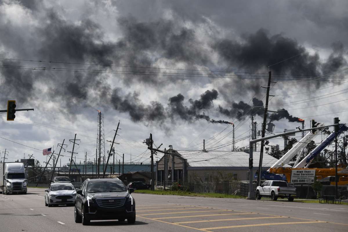 Trucks drive past leaning power lines and flaming smokestacks spewing black smoke into the air