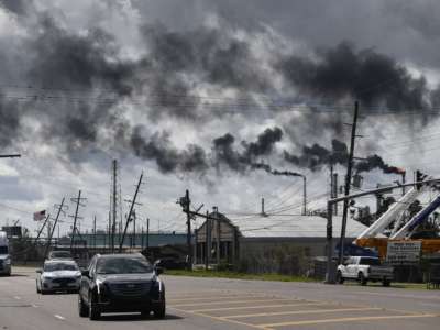 Trucks drive past leaning power lines and flaming smokestacks spewing black smoke into the air