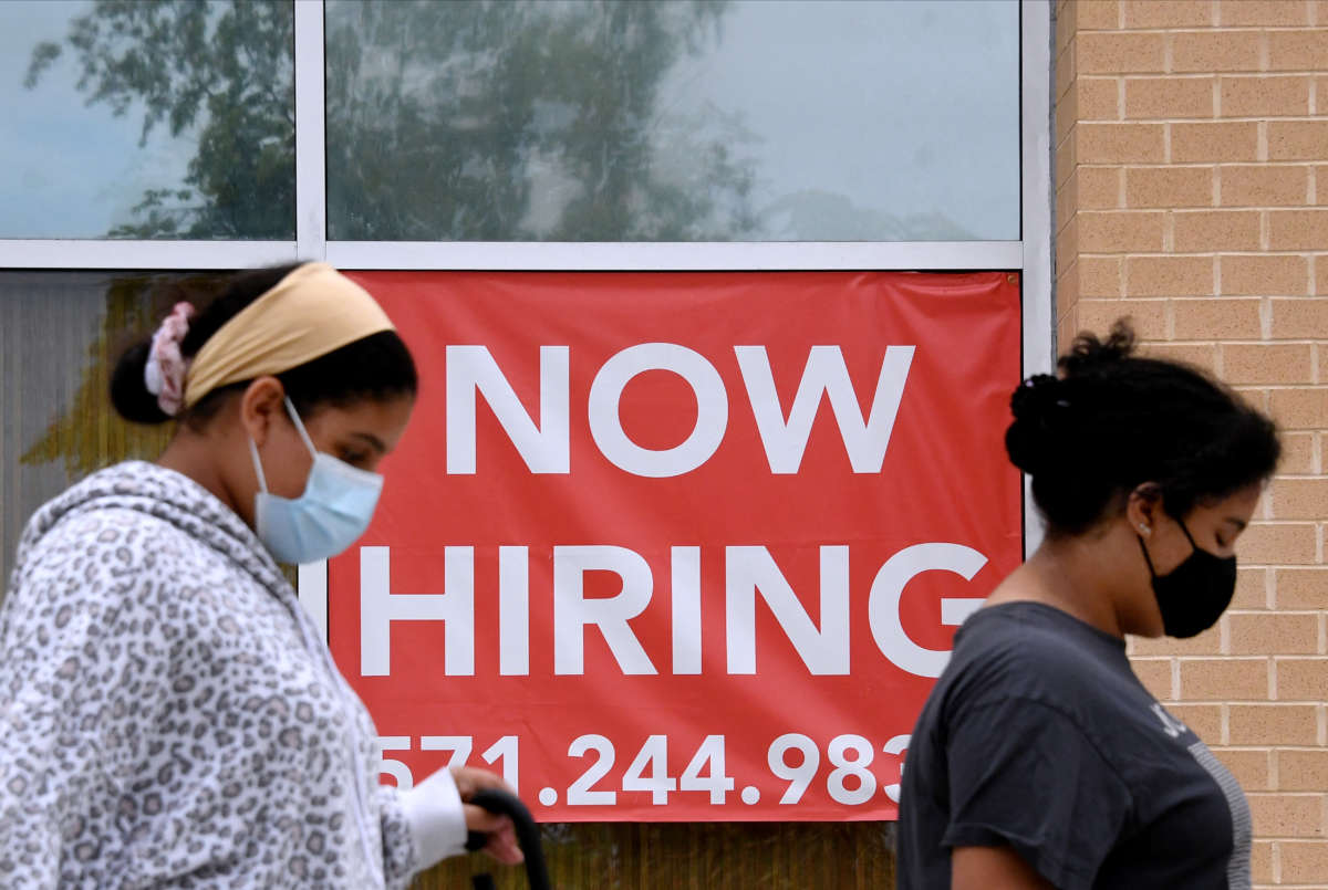 People walk by a "NOW HIRING" sign