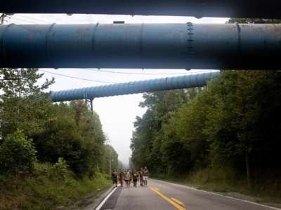 People march down an empty highway beneath what looks to be large pipelines