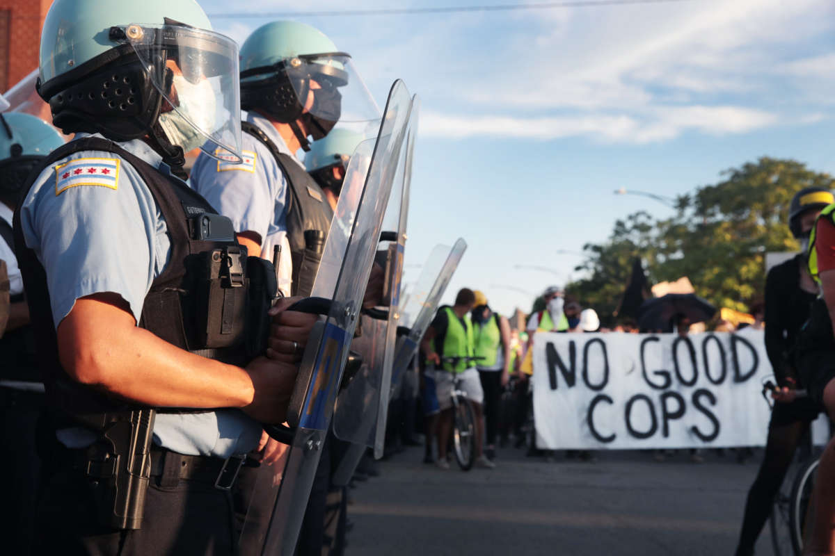 Police in riot gear stand by as they're accurately described by a banner in the background