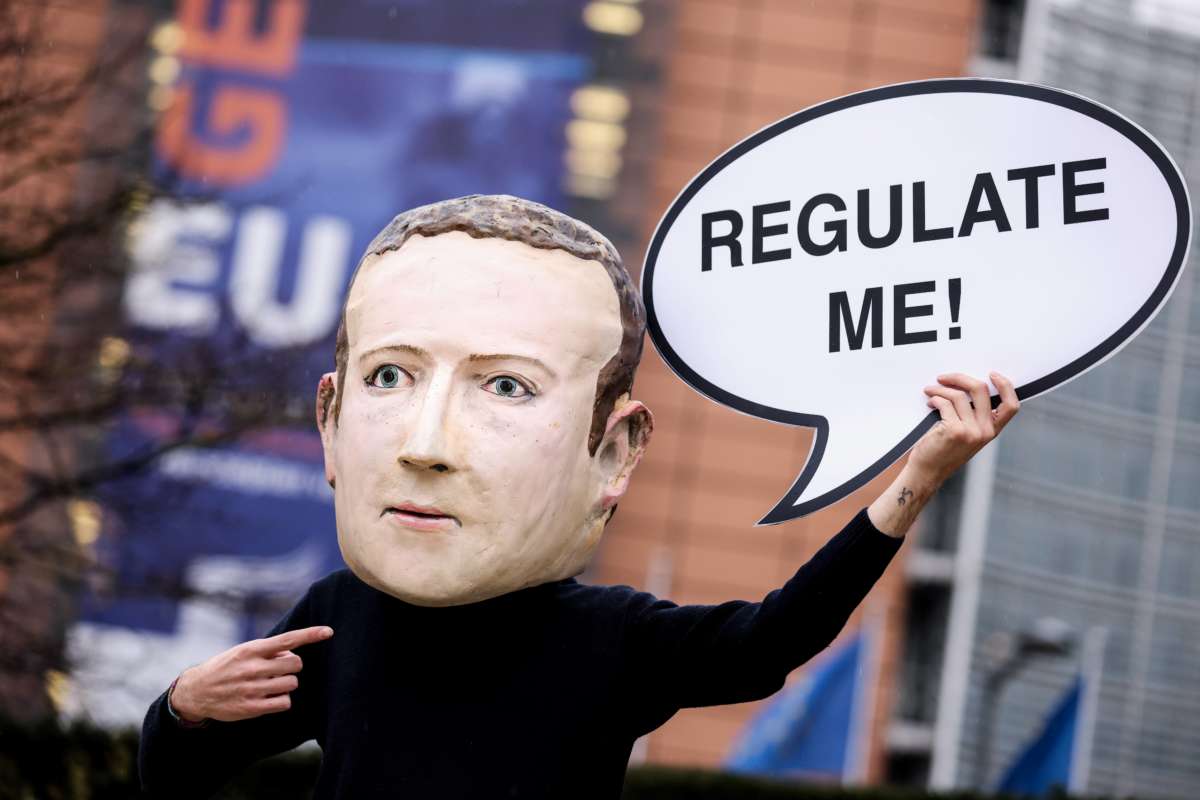 A protester in a deeply upsetting paper mache mask of Mark Zuckerberg holds a sign resembling a word bubble, with the text "REGULATE ME" written within