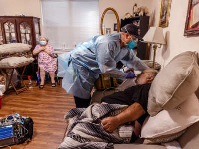 A man recieves medical care in his home while a woman in the background looks concerned