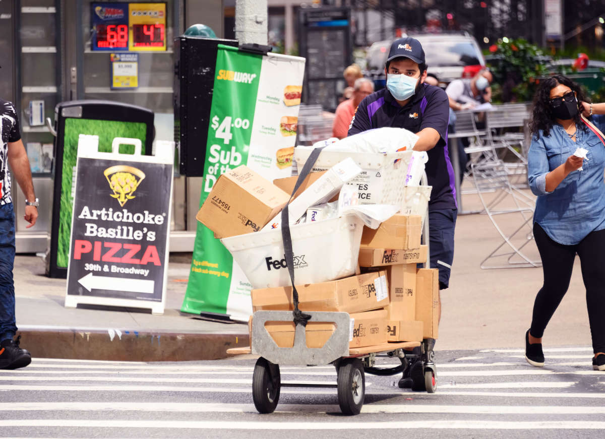 A masked fed-ex worker pushes a dolly stacked with packages