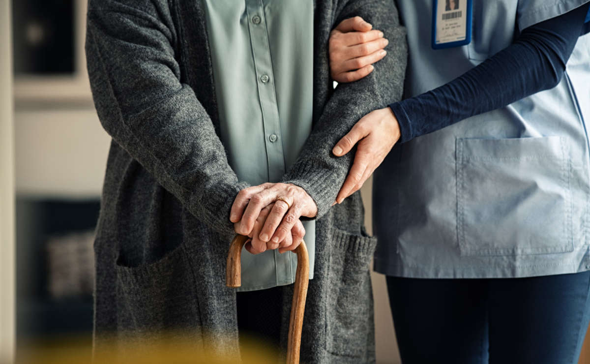 Elder woman using walking cane at nursing home with nurse holding hand for support.