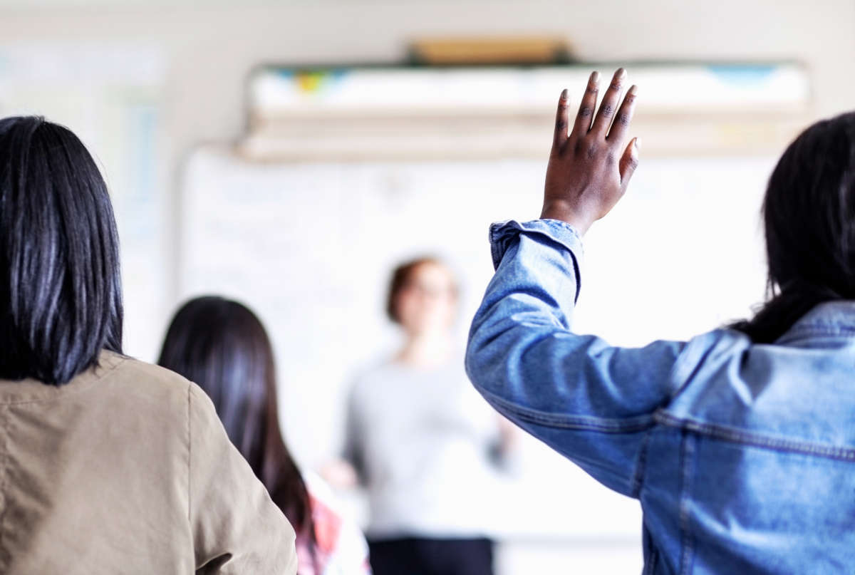 A Black student raises their hand as white teacher speaks, out of focus
