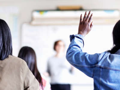 A Black student raises their hand as white teacher speaks, out of focus