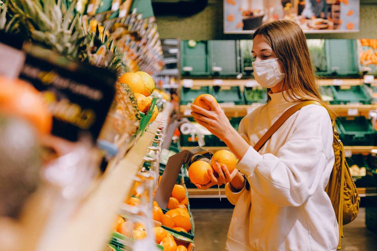 a masked woman holds several oranges in a grocery store.