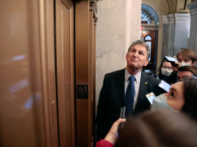 Joe manchin looks up at an elevator while surrounded by reporters