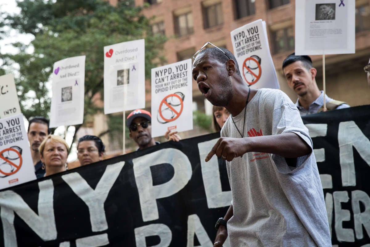 A activist speaks passionately as people behind him display signs decrying the NYPD's handling of the opoid crisis