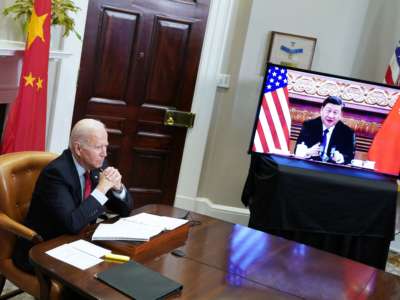 President Joe Biden folds his hands and looks forward as President Xi Jinping is seen speaking on a screen to Biden's left