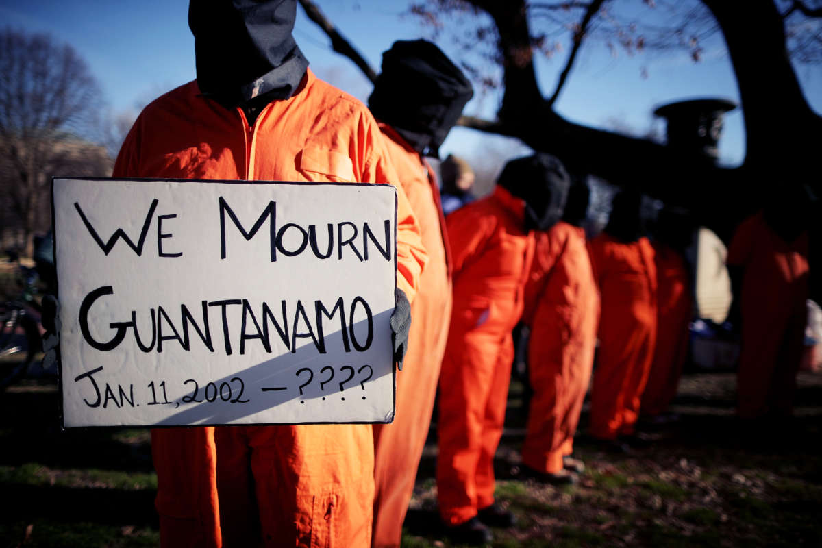 People in orange jumpsuits and hoods protest as one holds a sign reading "WE MOURN GUANTANAMO: JAN. 11, 2002 - ????"