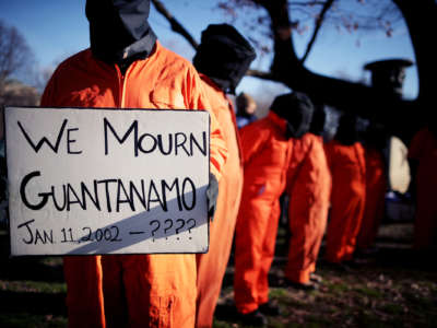 People in orange jumpsuits and hoods protest as one holds a sign reading "WE MOURN GUANTANAMO: JAN. 11, 2002 - ????"