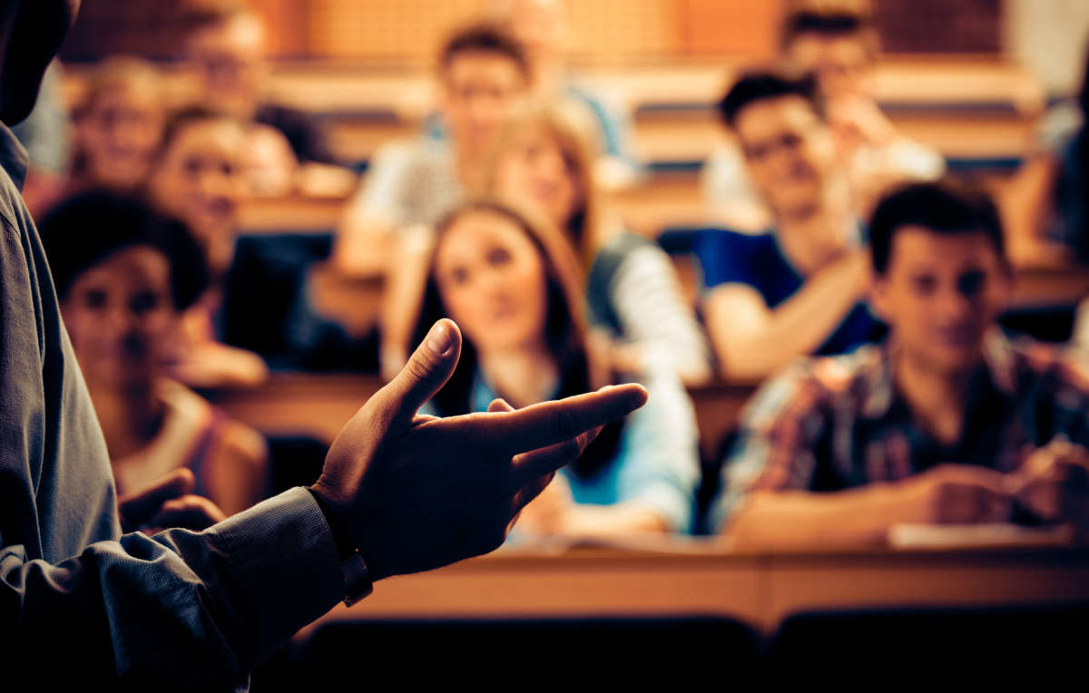 Large group of students sitting in the lecture hall at university and listening to their teacher, with focus on the professor's hand.