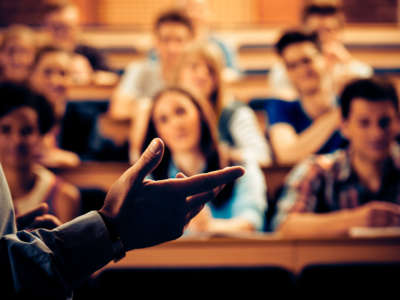 Large group of students sitting in the lecture hall at university and listening to their teacher, with focus on the professor's hand.