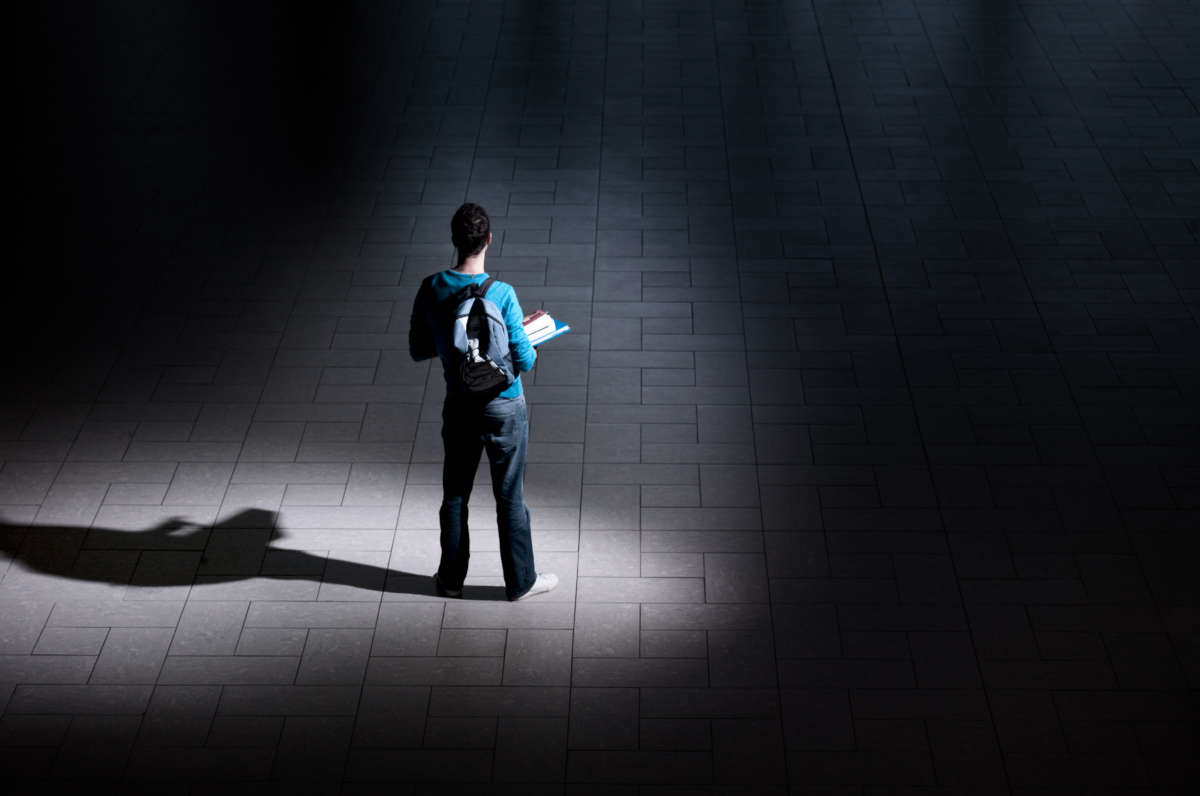 Student with backpack and books stands alone in large dark room