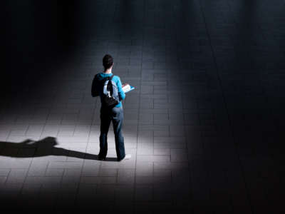 Student with backpack and books stands alone in large dark room