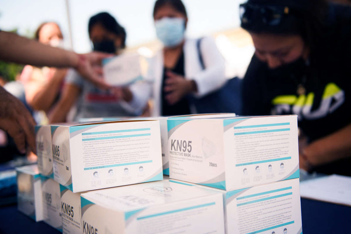 People fill out paperwork behind a stack of KN95 masks in the foreground