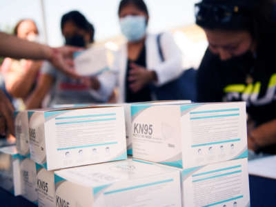 People fill out paperwork behind a stack of KN95 masks in the foreground