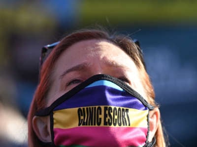 A protester wears a mask over their mouth and nose that reads "CLINIC ESCORT"