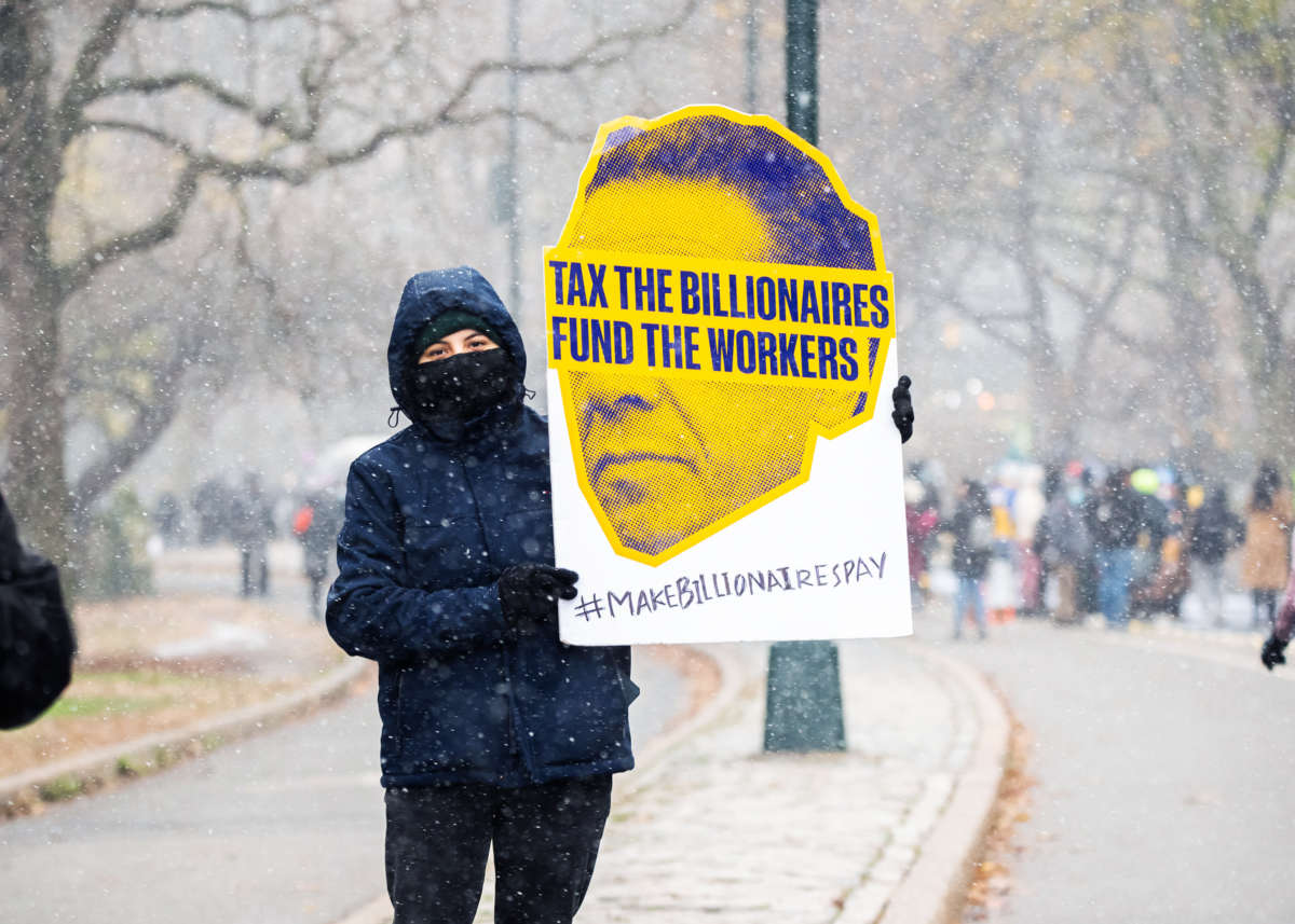 A masked protester holds a sign reading "TAX THE BILLIONAIRES; FUND THE WORKERS"