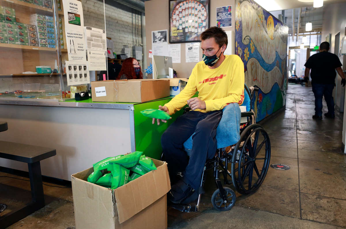 A person in a wheelchair tosses a green bag containing an overdose kit into a box filled with other, identical green bags