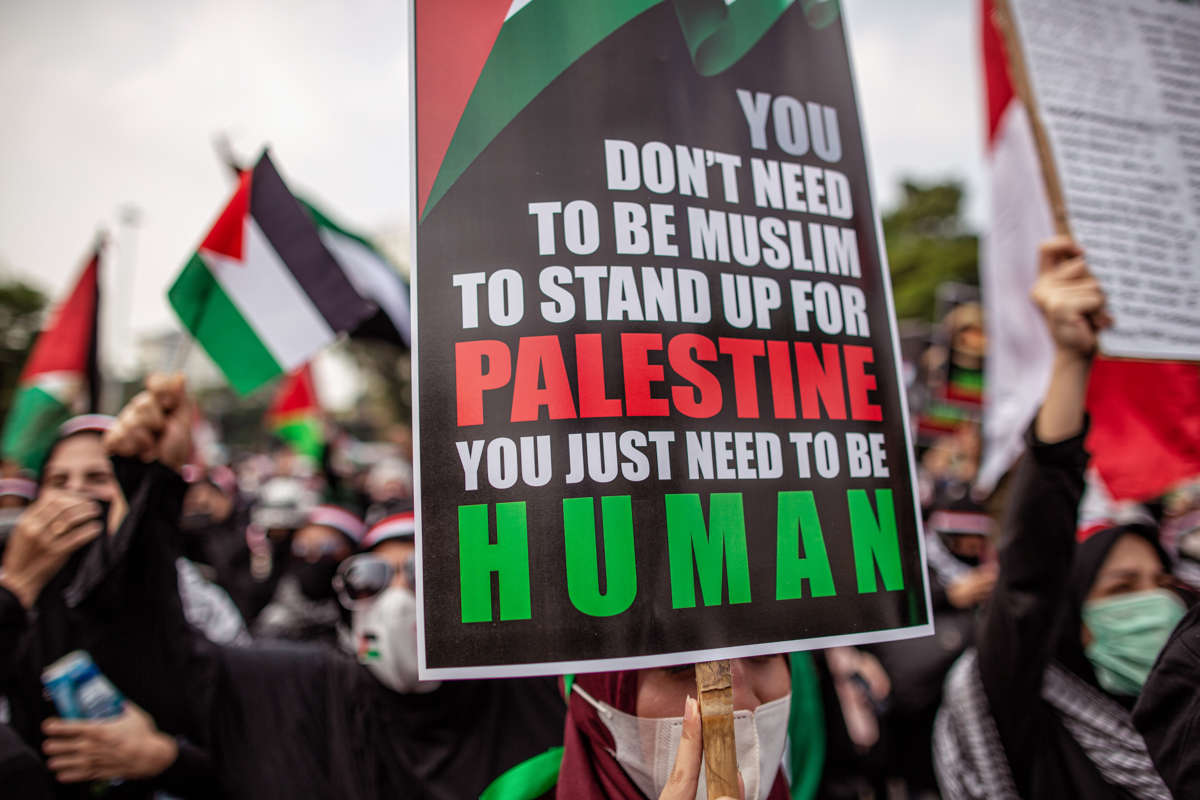 A protester holds a sign reading "YOU DON'T HAVE TO BE MUSLIM TO STAND UP FOR PALESTINE; YOU JUST NEED TO BE HUMAN"