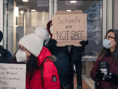 A masked protester holds up a sign reading "SCHOOLS ARE NOT SAFE" during an outdoor protest