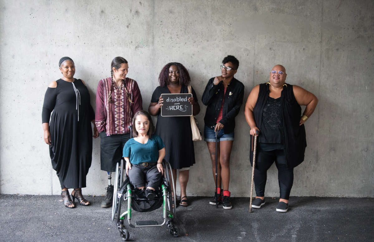 Six disabled people of color smile and pose in front of a concrete wall. Five people stand in the back, with the Black woman in the center holding up a chalkboard sign reading 'disabled and here.' A South Asian person in a wheelchair sits in front.