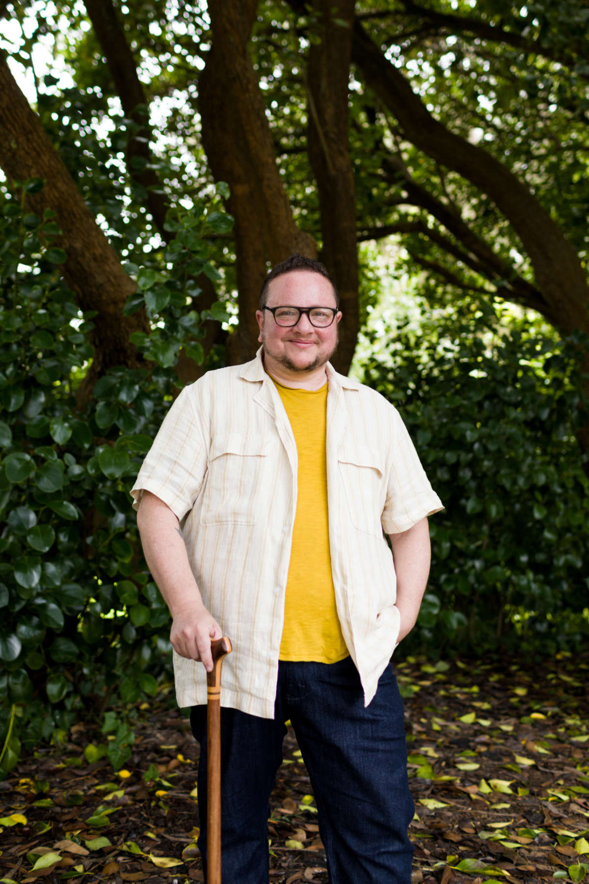 Rabbi Elliot Kukla, a white non-binary person, stands in front of a green background holding a wooden cane.