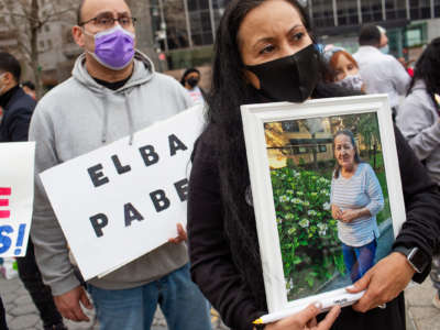 A masked woman at an outdoor protest holds a framed photo of an elderly loved one