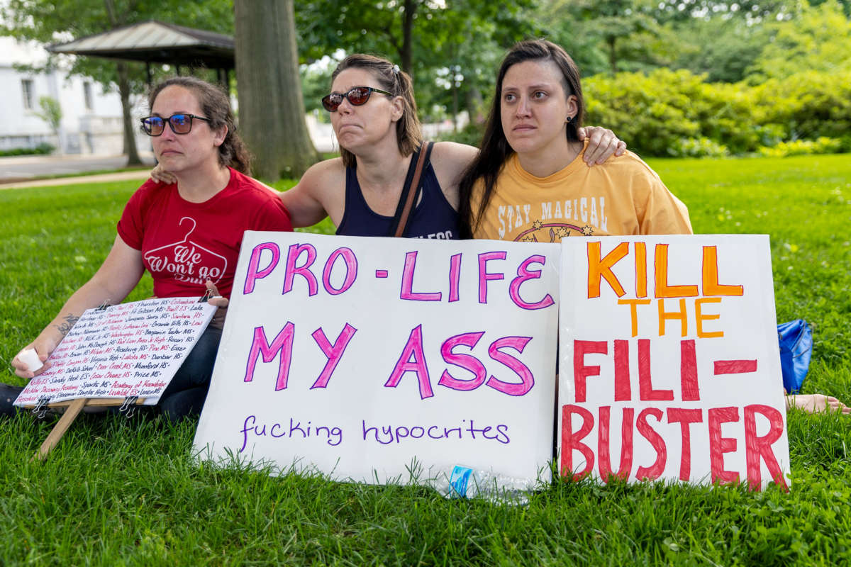 Three people embrace behind signs, one of which reads "PRO-LIFE MY ASS, FUCKING HYPOCRITES"