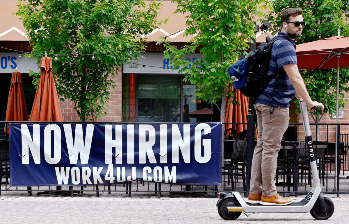 A person scooters past a "NOW HIRING" banner hung on a fence