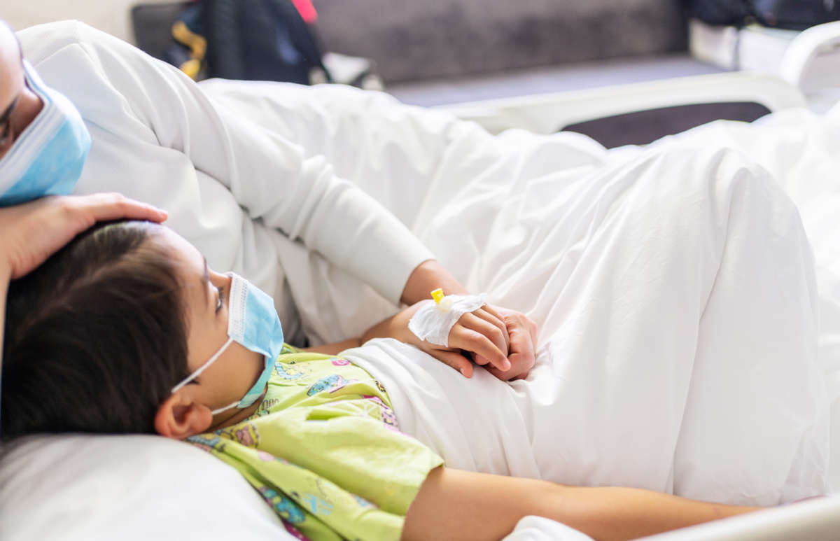 Mother with protective face mask holding son's hand while lying on the hospital bed with him.