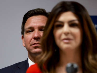 Gov. Ron Desantis lurks behind his wife as she speaks at a podium