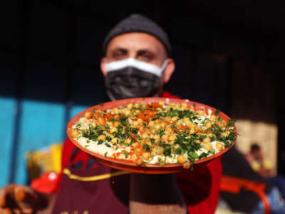 A masked man holds a plate of chickpea-topped hummus