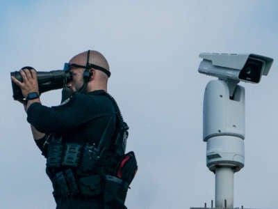 A Secret Service officer looks through huge binoculars while positioned on a roof