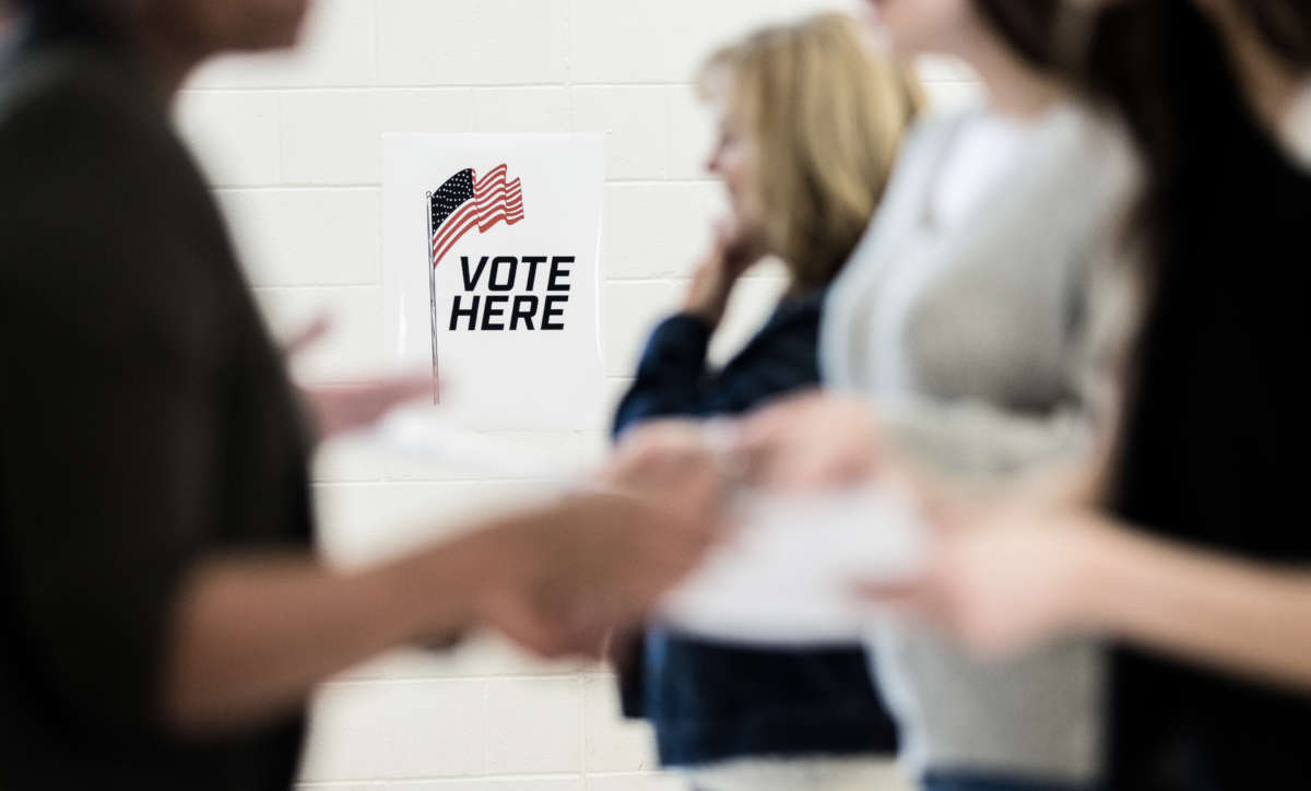 Group of voters, out of focus in foreground, in front of sign that reads 'Vote Here' at polling location