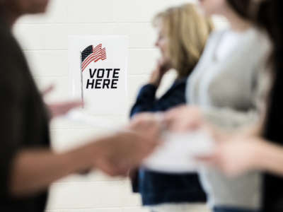 Group of voters, out of focus in foreground, in front of sign that reads 'Vote Here' at polling location