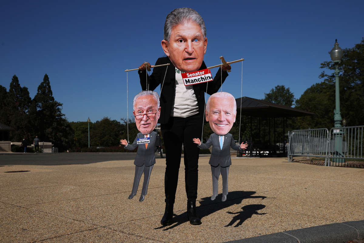 A protester dressed in a blazer and oversized print-out mask of Sen. Joe Manchin's head puppets effigies of Sen. Chuck Schumer and President Joseph Robinette Biden