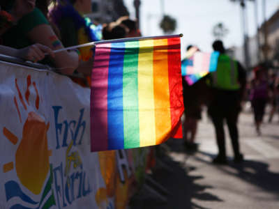 The sun shines through a gay pride flag held by a person participating in a Pride parade