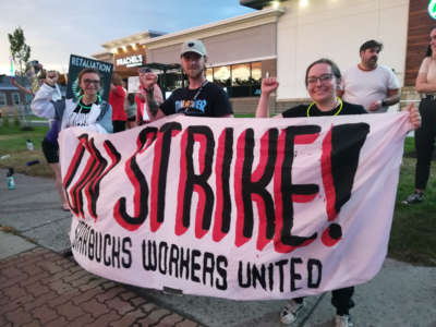 Workers raise their fists while holding a banner reading "ON STRIKE; STARBUCKS WORKERS UNITED"