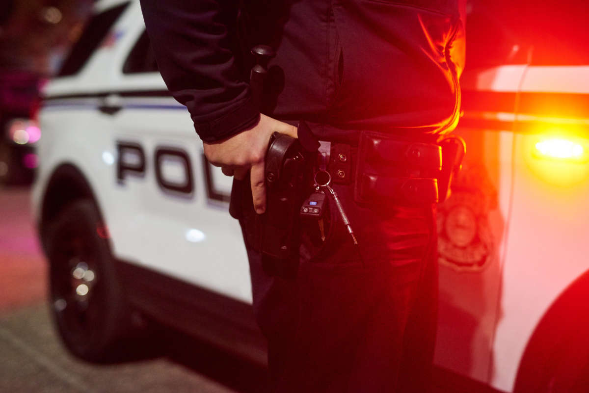 A police officer holds his gun in its holster while standing in front of police car